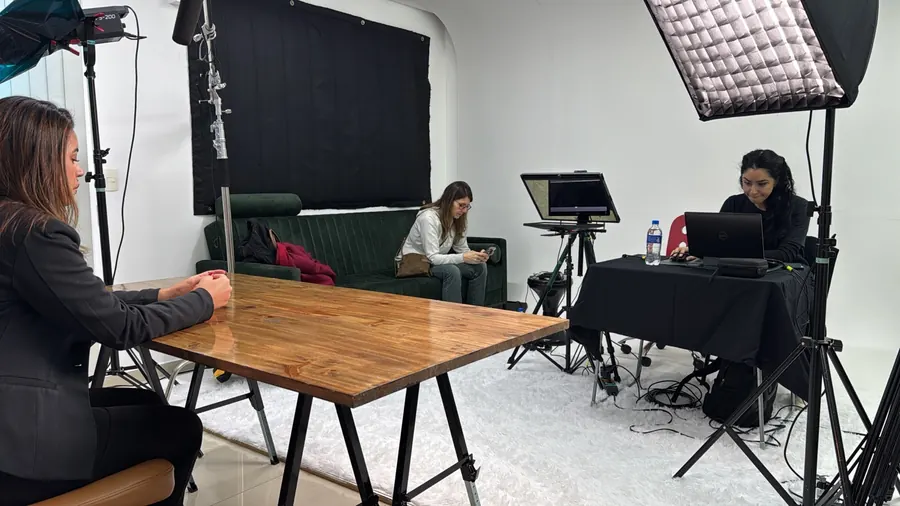 Gravação em andamento no Estúdio Gigante, com mesa de madeira, teleprompter, equipe operando notebook e sofá verde ao fundo